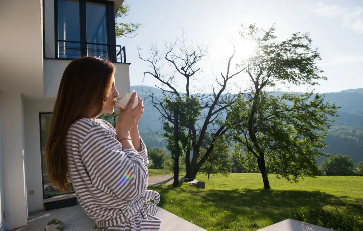 A woman with a cup of coffee looking out from a terrace over a green valley